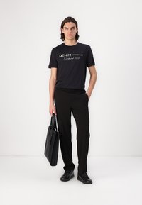 Juodas marškinėlis su balta inscription "DENIM TOM TAILOR Established 2007," derinamas su juodomis kelnėmis ir juoda rankine.
