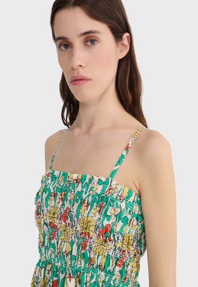 Jonge vrouw met lang bruin haar, gekleed in een mouwloze jurk met een groen-wit bloemenpatroon en dunne bandjes, tegen een effen achtergrond.