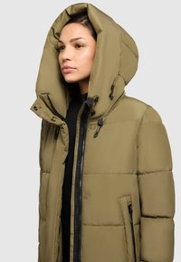 Olīvu zaļš pūšu jaka ar lielu, regulējamu kapuci, augstu apkakli un rāvējslēdzēja kabatām. Mīksta, pīlāru struktūra ar elegantu dizainu.