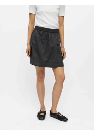 Personne debout devant un fond uni, portant une chemise blanche à manches courtes, une jupe noire longueur genou avec poches et des chaussures plates noires.