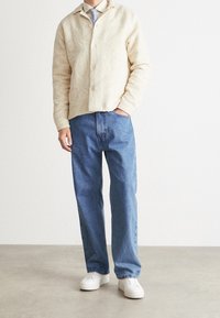 Beige mönstrad jacka med krage, blå raka jeans och vita sneakers, vilket visar en avslappnad outfit med en avslappnad passform.