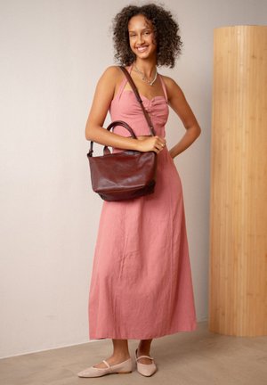 Glunderende vrouw in een mouwloze roze jurk met een zilveren halsketting, die een grote bruine leren handtas vasthoudt en binnen staat.