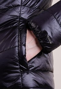 Veste rembourrée noire avec texture matelassée, poches latérales et poignets élastiques. Comprend une étiquette de marque sur la manche.