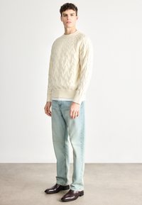 Krém színű, kötött pulóver bordázott mandzsettákkal és szegéllyel, világoskék farmerrel és sötétbarna bőrcsizmával párosítva. A modell sima háttér előtt áll.