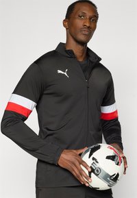 Μαύρο αθλητικό μπουφάν με κόκκινες και λευκές ριγέ λεπτομέρειες, ελαφρύ υλικό, κλείσιμο με φερμουάρ. Το μοντέλο κρατάει μια ποδοσφαιρική μπάλα με σχέδιο.