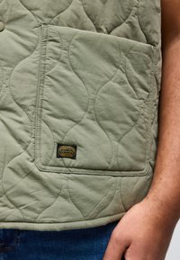 Τσεπάκι σε πράσινο ελιάς με επένδυση, με κίτρινη ετικέτα ρούχων Jack & Jones, σε άτομο που φοράει μπλε τζιν και έχει το χέρι του κοντά.