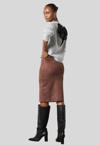 Femme portant un pull en peluche gris clair avec un détail de nœud noir dans le dos, une jupe en daim marron et des bottes noires à talons hauts jusqu'au genou, se tenant de profil.