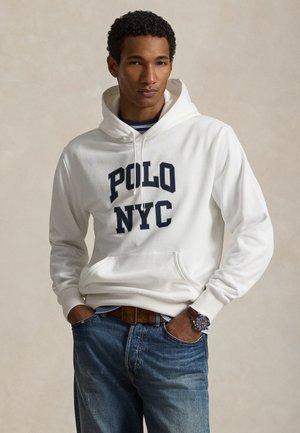Νέος άνδρας που φοράει λευκό φούτερ "POLO NYC", μπλε τζιν, καφέ ζώνη και ένα ρολόι χειρός, στέκεται με τα χέρια στις τσέπες.