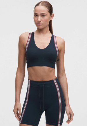 Vrouw die een marineblauwe sportbeha draagt en bijpassende hoge taille fietsbroek met rode en witte zijstrepen tegen een effen achtergrond.