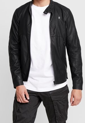 Mies pukeutuneena mustaan vetoketjulliseen nahkatakkiin, valkoiseen t-paitaan ja mustiin cargo-housuihin, joissa on vetoketju- ja läppätaskut.