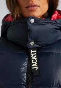 Marinblå vadderad jacka med hög krage och röd foder. Har en silversnapsknapp och en framträdande svart etikett med texten "JACK1T."