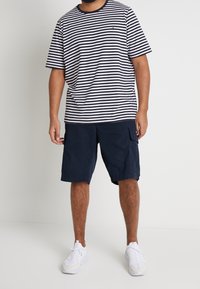 Muž ve slušivém tričku s černobílými horizontálními pruhy, námořnicky modrých cargo šortkách a bílých teniskách, stojící proti jednoduchému bílému pozadí.