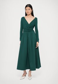 Φόρεμα μακρυμάνικο σε βαθύ πράσινο χρώμα με V-λαιμόκοψη, σούρες στη μέση και ρευστή σιλουέτα. Λείο ύφασμα με υφή, φορεμένο με λευκά τακούνια.