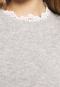 Pull en tricot gris avec un col rond, présentant une bordure en dentelle blanche qui dépasse au niveau du col. Texture douce, motif côtelé.