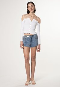 Crop top bianco a maniche lunghe con chiusura a bottone e design scollato, abbinato a pantaloncini di jeans blu sfilacciati. Sandali bianchi con cinturini.