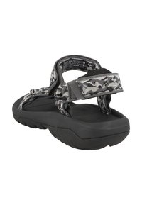 Musta sandaali, jossa on teksturoitu kumipohja, varustettuna säädettävillä harmailla kuviollisilla hihnoilla ja pehmeällä nilkatuesta.