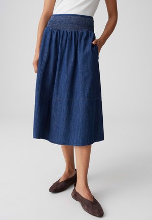 Frau trägt einen hoch taillierten, dunkelblauen Jeans-Midirock mit seitlichen Taschen und braunen flachen Schuhen, steht vor einem einfarbigen Hintergrund.