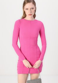 Vaaleanpunainen pitkähihaisuus mini-mekko, jossa on teksturoitu rypytetty muotoilu, istuva siluetti ja pyöreä kaula-aukko. Malli pitää kädessään pientä hopeista laukkua.