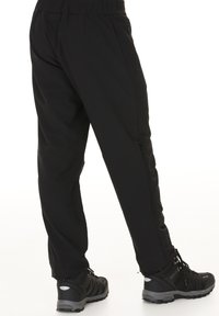 Mustat pehmeät housut elastisella vyötäröllä, sivussa vetoketjutasku ja kapeneva lahkeen muotoilu, yhdistettynä mustiin vaelluskengiin.