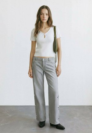 Žena stojaca, oblečená v bielom tričku s krátkym rukávom, svetlo sivé široké nohavice so zipsami na boku, čierne špicaté čižmy a zelená kabelka na rameno.