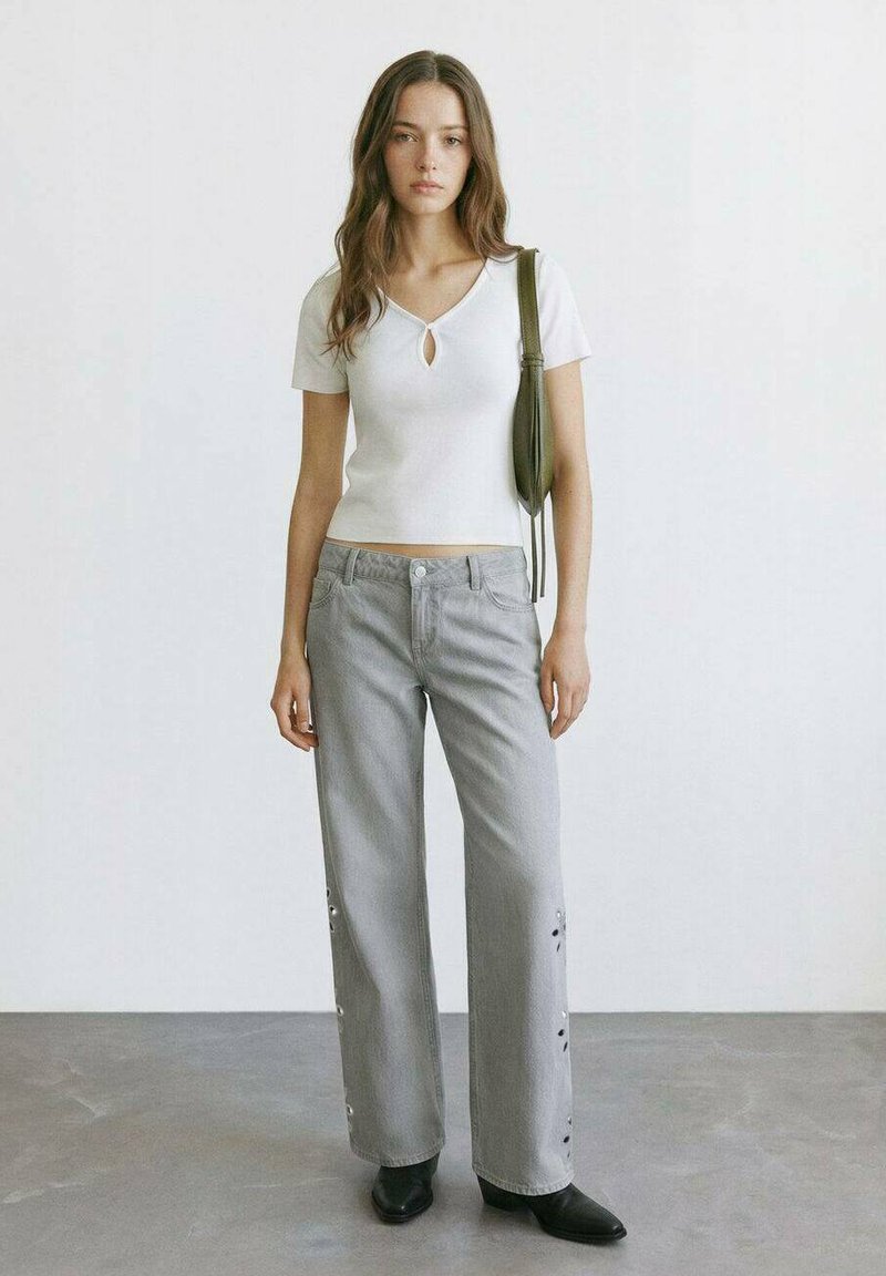 Donna in piedi che indossa una maglia bianca a maniche corte, pantaloni ampi grigio chiaro con occhielli laterali, stivali neri con punta e una borsa verde a tracolla.