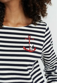 Gestreiftes langärmliges Shirt in Marineblau und Weiß mit einem roten gestickten Anker, auf dem ein Snoopy-Charakter steht. Aus weichem Baumwollstoff.