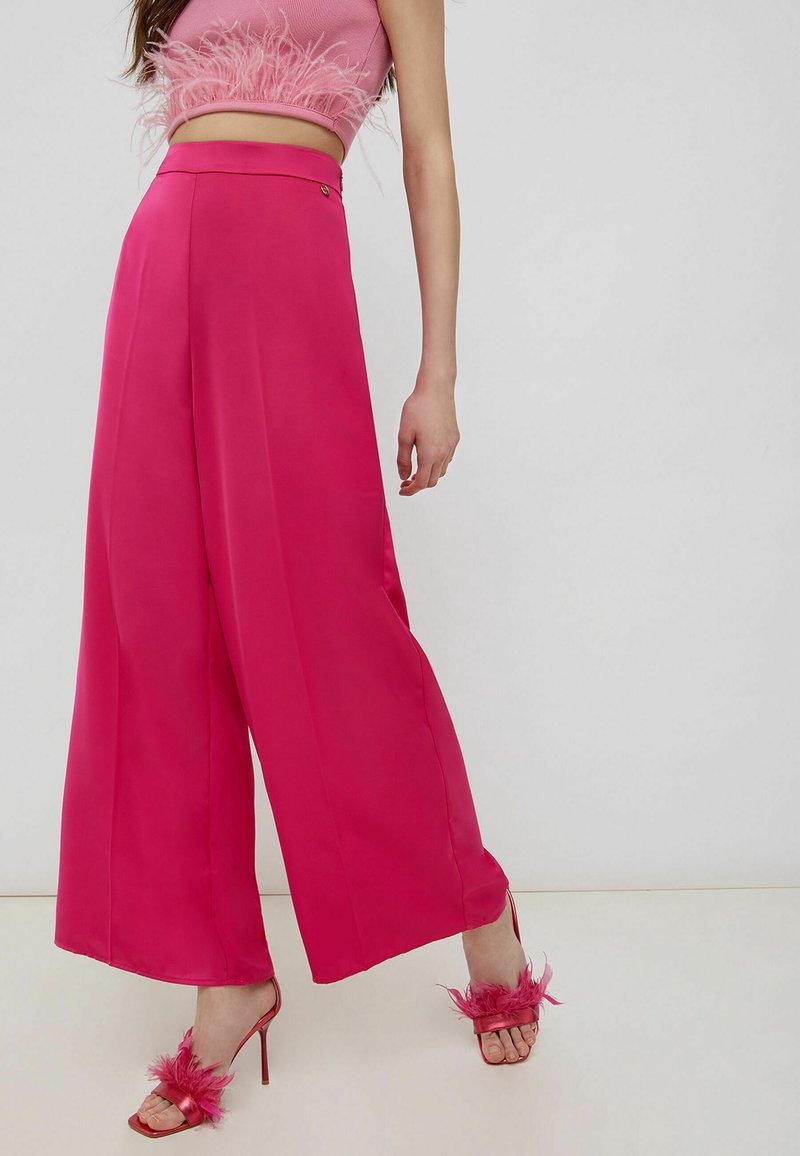 Vrouw in een felroze wijde broek, bijpassende croptop met verenrand en roze gevederde hoge hakken voor een effen achtergrond.