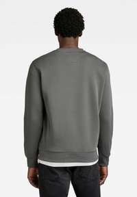 Tmavě šedá mikina z měkké látky, s kulatým výstřihem, žebrovanými manžetami a malou čtvercovou nášivkou na zádech.