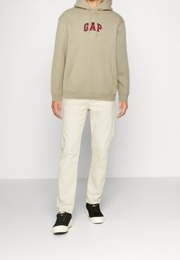 Sudadera con capucha beige con logo rojo "GAP", bolsillo delantero, combinada con pantalones blanco roto y zapatillas negras. Diseño simple y ajuste casual.