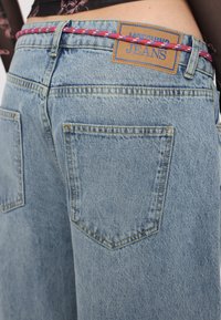 Hellblaue Denim-Jeans mit einem braunen Lederschild, auf dem "Moschino Jeans" steht. Sie haben einen pinkfarbenen, gemusterten Kordelzug und zwei Gesäßtaschen.