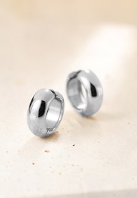 Coppia di anelli in argento lucido, con design curvo e finitura lucidata, adagiati su una superficie leggera e texture.