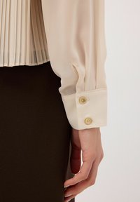 Blusa plissettata beige chiaro con texture liscia e maniche con polsini abbottonati. Bottoni dorati aggiungono dettaglio ai polsi. Gonna marrone scuro sotto.
