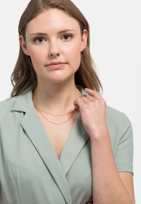 Náhrdelník z ružového zlata, vrstvený, s malými perličkami, predvedený na modeli v svetlozelenom blúzke. Jednoduchý, jemný dizajn s hladkou textúrou.