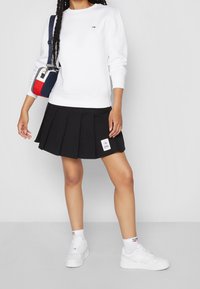 Felpa bianca con un piccolo logo, gonna plissettata nera e sneakers bianche. Include una piccola borsa a tracolla con dettagli rossi, blu navy e bianchi.