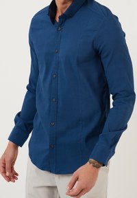 Niebieska koszula z długim rękawem, wykonana z teksturowanej tkaniny, z kołnierzykiem w kształcie ostrokońca oraz mankietami zapinanymi na guziki, z ciemnymi guzikami wzdłuż listwy guzika.