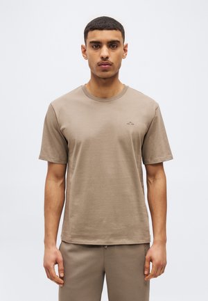 Mladý muž oblečený v obyčajnom béžovom tričku s krátkym rukávom a okrúhlym výstrihom a nohaviciach rovnakej farby, stojaci pred jednoduchým svetlým pozadím.