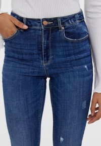 Siniset korkeavyötäröiset farkut, joissa on istuva muotoilu. Ominaisuuksiin kuuluu kaksi etutaskua, kulutettuja yksityiskohtia ja messinkinen nappisulku.