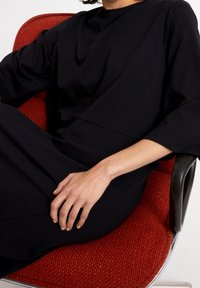 Czarna sukienka o luźnym kroju, z długimi rękawami i gładką fakturą. Ręka modelki spoczywa na teksturowanym czerwonym fotelu.