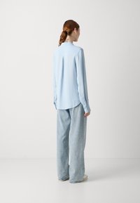 Camicia azzurro chiaro a maniche lunghe con texture plissettata, colletto e polsini abbottonati. Abbinata a jeans blu chiaro a vestibilità ampia.
