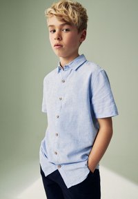 Lyseblå kortærmet skjorte af linned med knapper foran i træ og en spredt krave, kombineret med mørke bukser.