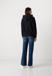 Juodas gobtuvas su gobtuvu, derinamas su mėlynomis plačiomis džinsomis ir baltomis sportbačiais. Lygus audinys, laisvas siluetas ir minimalistiškas dizainas.