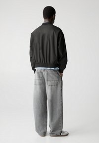 Sort bomberjakke med elastiske manchetter og talje, parret med lysegrå brede jeans med to baglommer. Glatte teksturer overalt.