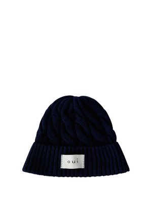 Bonnet tricoté bleu foncé avec bord-côtes et une étiquette rectangulaire blanche marquée "oui" à l'avant.