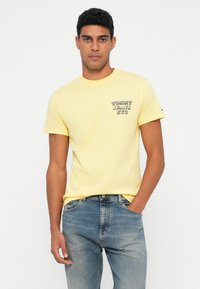 Noor mees, seljas kollane Tommy Jeans NYC t-särk ja sinised teksad, seisab ühe käega taskus, taustaks ühtlane valge sein.