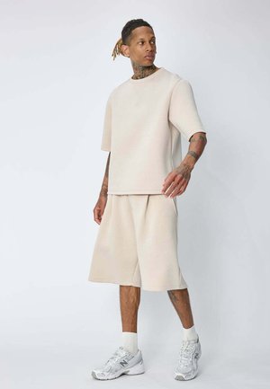 Homme tatoué debout en t-shirt oversize écru et short large assorti, chaussettes blanches et baskets, sur fond clair uni.