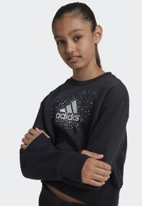 Sudadera negra de manga larga, con puños acanalados y cuello redondo. Presenta el logo de adidas en blanco y un patrón de puntos dispersos como detalle.