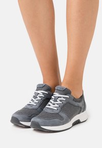 Graue Sportschuhe mit Mesh-Einsätzen, Wildlederakzenten, weißen Schnürsenkeln und einer dicken weißen Sohle. Das Design ist ein Low-Top-Modell mit gepolstertem Kragen.