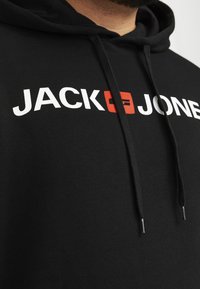 Sort hættejakke med snor, prydet med store hvide og røde bogstaver "JACK & JONES." Fremstillet af bomuldsstof med en glat overflade. Minimalistisk design.