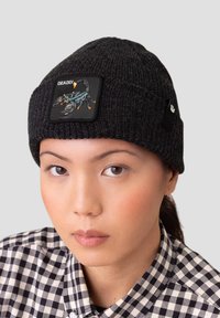 Bonnet en tricot noir avec un patch carré présentant un design de scorpion en bleu et orange. Texte "MORTEL" sur le patch.