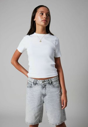 Ung kvinde stående med hånden på hoften, iført hvid kortærmet skjorte, acid-wash denimshorts og en guldkæde med vedhæng.